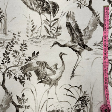 Ljust mönstrat tyg Cordelia i 100 % bomull med illustrerade tranor, träd och växter i mjuka grå och beige toner, fotograferat plant med måttband för skala.