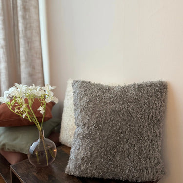 Grå kudde i mjukt bouclétyg Dolly i 100% polyester, placerad på träbord bredvid vas med vita blommor. Dekorativ kudde för soffa eller fåtölj i varm och ombonad inredningsmiljö.