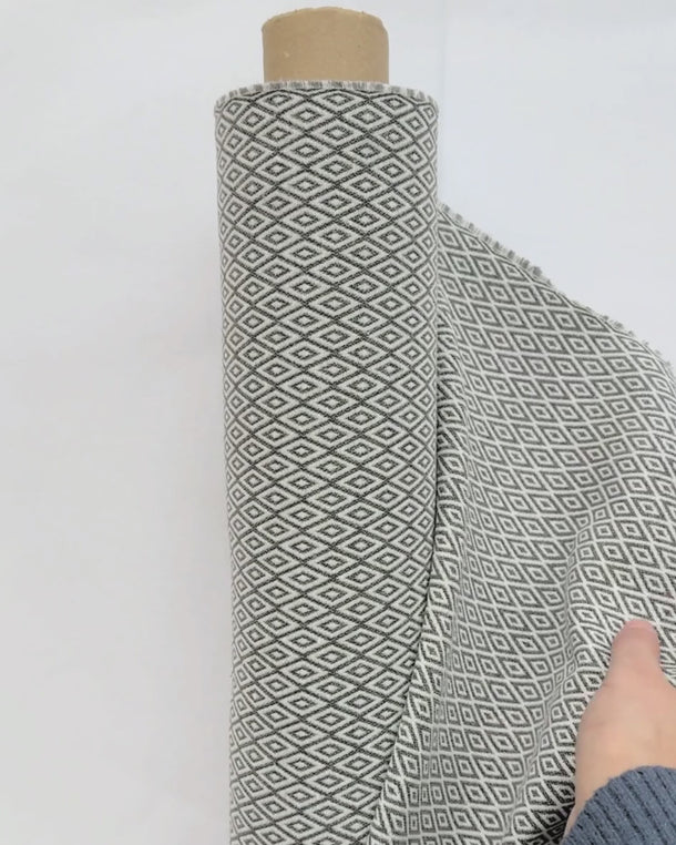 Video där en hand lyfter möbeltyget Saga i grå ton med små geometriska mönster för att visa tygets struktur, väv och tjocklek.