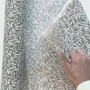 Hand som visar strukturen på det mönstrade inredningstyget Ramas med kvistar och blad, perfekt för kuddar och dukar.