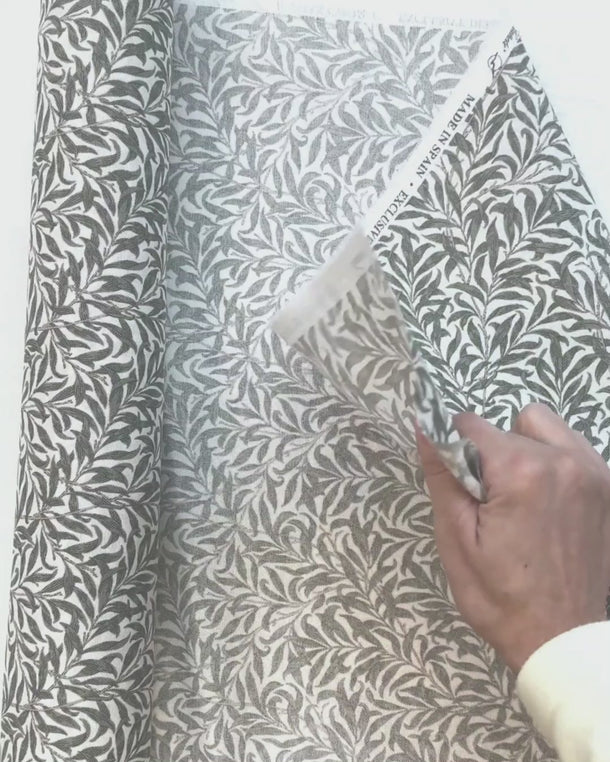 Hand som visar strukturen på det mönstrade inredningstyget Ramas med kvistar och blad, perfekt för kuddar och dukar.